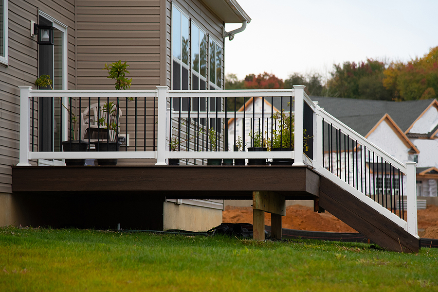 A dark-brown stained deck with white railings protruding from a beige house atop green grass.