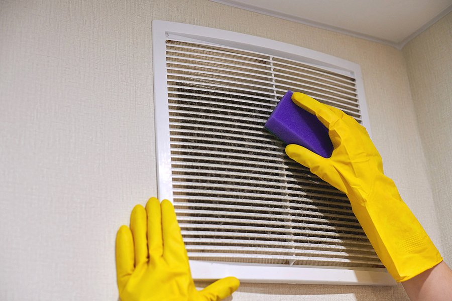 Yellow gloved hands scrubbing a vent cover with a blue sponge.