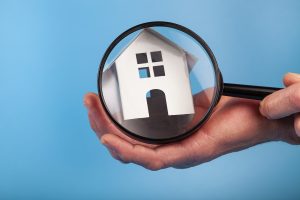 Hand holds house replica under a magnifying glass to signify concept of a home inspection.